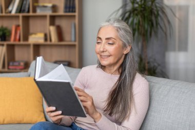 Smiling pretty caucasian elderly gray-haired female resting, relaxing and reading book in living room interior. Hobbies in free time at comfort home and literary and cozy during covid-19 quarantine