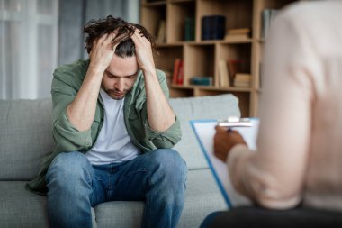 Depressed despaired millennial european male suffers from mental problems, listens to black female psychologist in clinic interior. Psychological medical care, disease treatment and support, therapy
