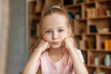 Sad bored pretty schoolgirl pupil studying at home, resting head on hand and looking at camera. Overwork, problems with remote education, boring lessons concept