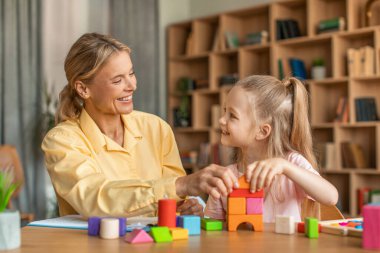 Friendly female child development specialist having session with cheerful little girl, sitting at table and playing with colorful wooden bricks, smiling at each other