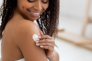Body Care Concept. Smiling Beautiful Black Woman Applying Moisturizing Lotion On Shoulder, Happy African American Female Making Beauty Treatments While Standing Wrapped In Towel After Bath, Closeup