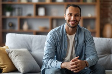 Portrait Of Handsome Young Black Man Posing In Home Interior, Positive African American Guy Sitting On Comfortable Sofa And Smiling At Camera, Enjoying Domestic Leisure, Closeup Shot With Copy Space