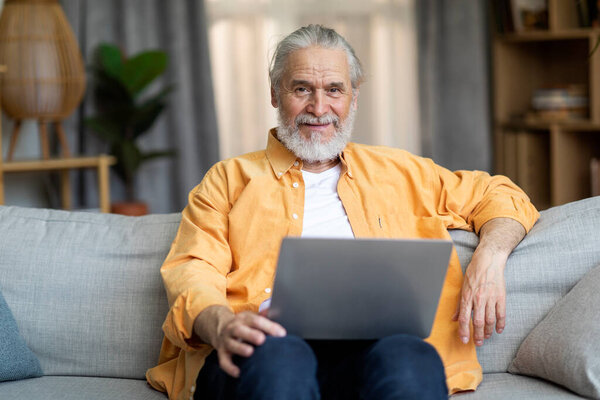 Happy grandfather stylish grey-haired senior man websurfing while resting at home, sitting on couch and using modern laptop, living room interior, looking at camera and smiling, copy space