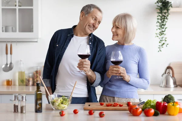 Loving elderly spouses drinking red wine while cooking together at home, happy senior husband and wife preparing dinner, having conversation, kitchen interior, copy space. Marriage, relationships