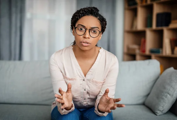 Serious young black woman doctor in glasses speaks and gesticulates, looking at camera in office clinic interior. Online support, medicine, psychological therapy, session and mental help remotely