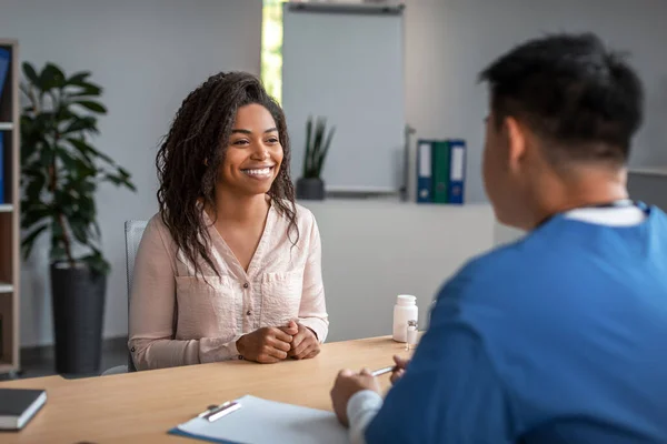 Happy pretty young african american woman patient in consultation with asian man doctor in uniform in office clinic interior. Health care, meeting with therapist and treat illness, medical service