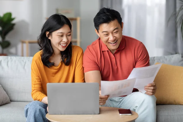 Happy asian spouses paying bills online while staying home, sitting on couch, using laptop, happy chinese man holding papers and banking on Internet, cheerful woman typing on keyboard, copy space