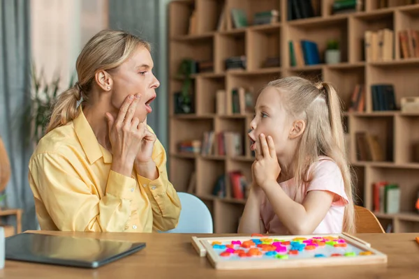 Professional woman speech therapist studying together with pretty little girl, learning practice pronunciation exercises with child at office, touching faces and grimacing, sitting at desk