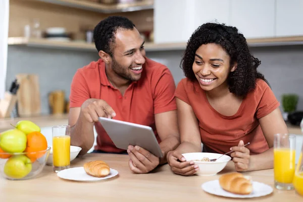 Laughing young african american couple have breakfast, enjoy chatting, watching video in tablet and good morning in kitchen interior. New app and online call, meeting remote, love and relationships