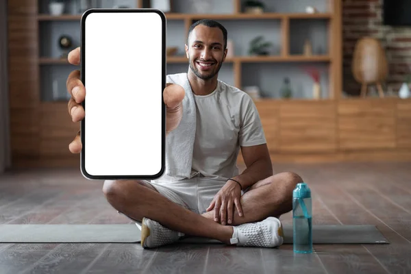 Fitness App. Happy Black Man Training At Home And Showing Big Blank Smartphone With White Screen At Camera, Smiling African American Guy Demonstrating Copy Space For Mobile Design, Mockup, Collage