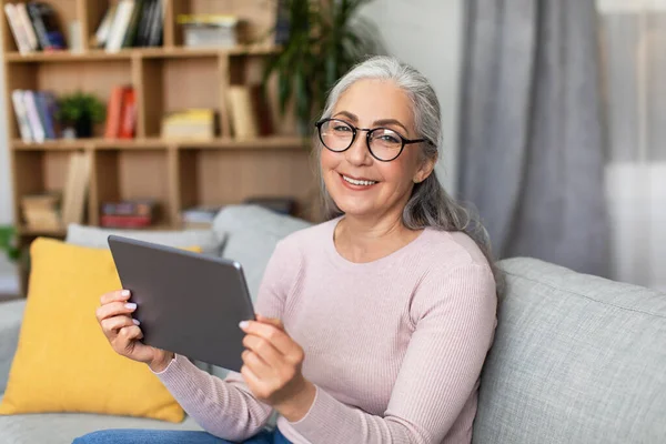 Smiling mature european gray-haired woman in glasses hold tablet, chatting, watch video in living room interior. Social networks, blog, video call and new app, health care during covid-19 pandemic
