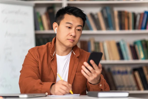 Asian Middle Aged Man Using Smartphone Learning Or Teaching Online Wearing Earbuds Sitting In Library Indoor. Educational Application, E-Learning Concept. Selective Focus