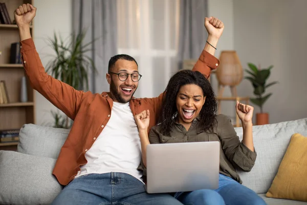 Joyful Black Couple Shaking Fists Looking At Laptop Computer Gesturing Yes Celebrating Great News Sitting On Couch At Home. Victory And Success Celebration Concept