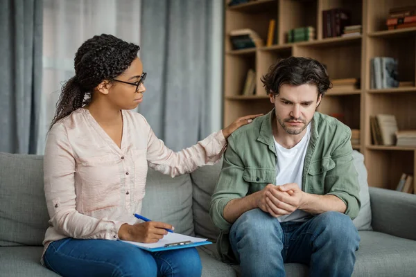 Serious young black female doctor calms european guy, client suffering from depression in office clinic interior. Medicine, psychological therapy, session with professional, mental help and emotions