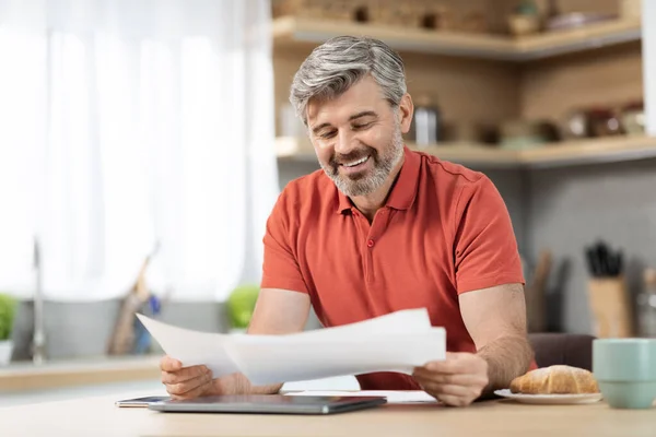 Handsome middle aged businessman working from home, happy grey-haired man having breakfast at kitchen, reading papers and smiling, copy space. Remote job, online business concept
