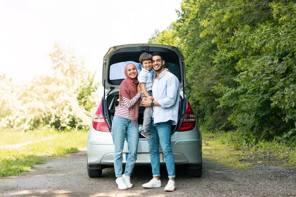 Rest outdoor from road, family journey at summer. Satisfied millennial arab man and lady in hijab holding baby near car with open trunk, copy space. Trip, travel together, vacation and people emotions