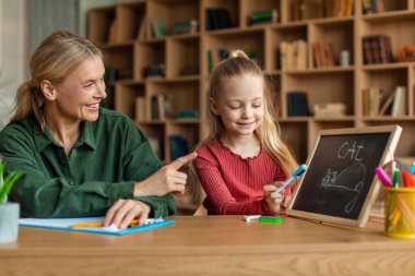 Cheerful female speech therapist teaching adorable little girl alphabet, child writing word on blackboard with chalk. Speech-language therapy for children concept
