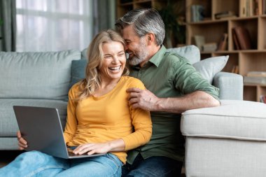 Happy Moments. Romantic Middle Aged Spouses Using Laptop And Bonding At Home, Cheerful Mature Husband And Wife Laughing And Chatting In Living Room Interior, Enjoying Spending Time Together