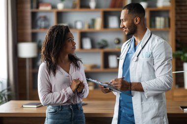 Friendly handsome middle eastern man doctor consulting young african american lady patient, doc giving recommendations, holding medical chart, standing next to workdesk at modern clinic