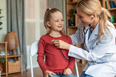 Female pediatrician visiting her little patient at home, woman in uniform listening to child patient breath with stethoscope, copy space. Happy kid looking at doc and smiling