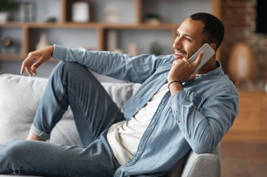Cheerful Black Guy Talking On Cellphone And Relaxing On Sofa At Home, Happy Young African American Man Having Phone Call With Friends While Resting On Sofa In Living Room, Side View, Closeup
