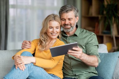 Happy Middle Aged Spouses Watching Videos On Digital Tablet Together While Resting On Comfortable Sofa In Living Room At Home, Smiling Mature Couple Enjoying Domestic Leisure, Closeup Shot