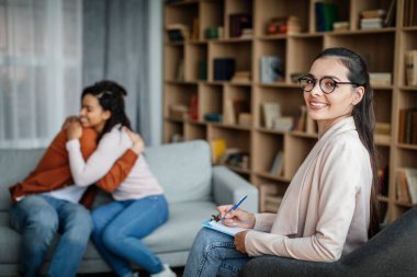 Glad young caucasian male hugging black female at meeting with doctor psychologist in office clinic interior. Family psychological therapy, consultation together, mental treatment and solving problems