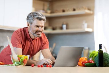Cheerful grey-haired middle aged handsome man in casual cooking alone at home, reading food blog, looking for nice recipe online, making vegetable salad, using laptop at kitchen, copy space