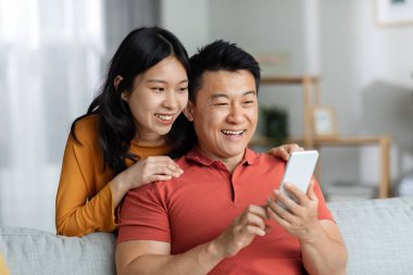 Cheerful chinese lovers having video chat with friends, positive asian couple making online party with family while staying together at home, using mobile phone, looking at gadget screen and smiling