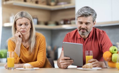 Suspicious blonde lady watching her handsome middle aged husband using digital tablet while having breakfast together, chatting with ladies on social media, kitchen interior, copy space