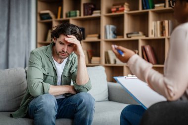 Unhappy depressed young european male patient listens to black woman psychologist in clinic interior. Psychological support, professional medical care, mental problems, disease treatment and help