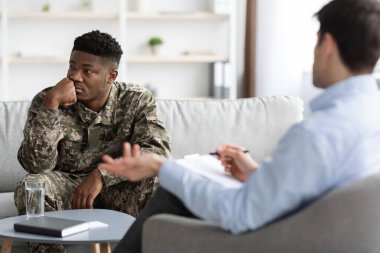 Depressed young black guy in camouflage uniform combat having therapy session with psychologist, sitting on sofa in clinic, looking aside while listening to doctor, copy space. PTSD for veterans