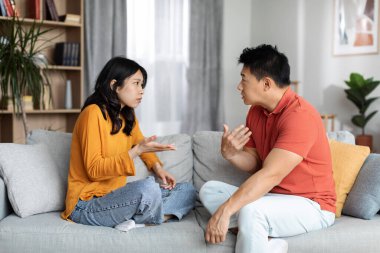 Emotional japanese lovers having quarrel at home, furious asian middle aged man and young woman sitting on sofa in front of each other, yelling and gesturing, experiencing difficulties in marriage