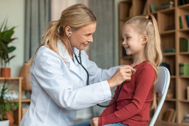 Happy little girl having checkup at pediatrician, pretty child sitting on chair and smiling while female doctor checking up her chest with stethoscope
