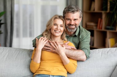 Portrait Of Smiling Middle Aged Spouses Posing In Home Interior, Romantic Mature Couple Embracing And Looking At Camera While Resting On Sofa In Cozy Living Room, Enjoying Time Together, Free Space