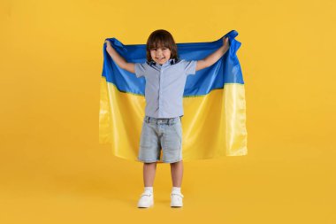 Little patriot. Happy adorable preteen boy posing wit Ukrainian flag and laughing, loving his motherland, yellow studio background