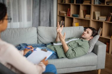 Sad young european guy patient lies on sofa and talks with black woman doctor in office clinic interior. Support, medicine, psychological therapy, session with professional, mental help and pressure