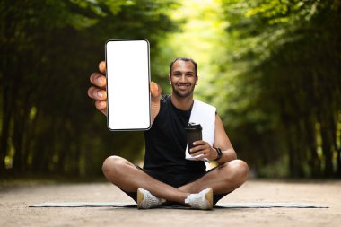 Cheerful young athletic african american man in sportswear training in park, showing brand new smartphone with white blank screen and smiling, recommending nice mobile fitness app for tracking, mockup