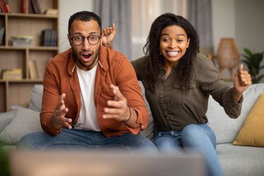 Emotional African American Spouses Shaking Fists In Joy Watching News On TV Sitting On Sofa At Home. Couple Celebrating Victory Of Favorite Team. Selective Focus
