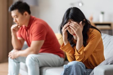 Upset chinese couple having fight at home, unhappy asian man and woman sitting on couch far from each other, looking down, thinking about divorce, cant solve conflict, breakup after quarrel