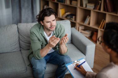Sad emotional young european man gesturing and talking to black female psychologist in clinic interior. Professional medical care, mental problems, treatment of depression, lifestyle and consultation