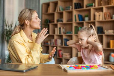 Unruly little girl closing ears with hands expressing negative emotions and looking at teacher, having private development lesson in cabinet. Woman shouting at child