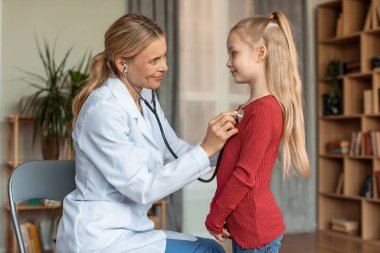 Cheerful female pediatrician listening girls heartbeat with stethoscope during checkup at home, pediatrist doctor checking childs heart and lungs, side view
