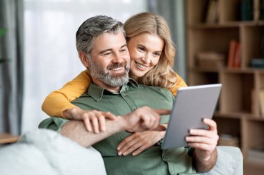 Happy Middle Aged Couple Using Digital Tablet While Relaxing In Living Room Together, Smiling Mature Man And Woman Shopping Online, Browsing Internet Or Checking New Application, Closeup Shot