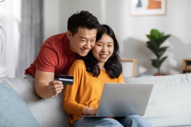 Smiling asian husband and wife embracing, using modern computer and plastic credit card, cheerful japanese family paying for goods and services online while staying home, copy space