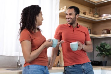 Cheerful millennial african american husband and wife in same t-shirts with cups enjoy drink and free time together in kitchen interior. Breakfast and good morning, coffee break, relationship and love