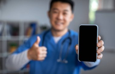 Smiling young asian nurse in uniform with stethoscope shows smartphone with blank screen and thumb up in office clinic interior, selective focus. Health care remote, app approval, gadget for medicine