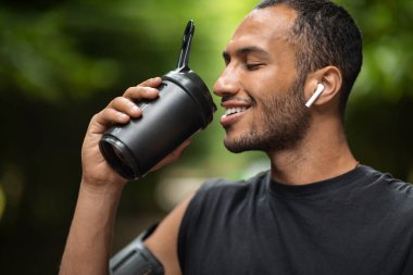Closeup of handsome young african american man drinking protein cocktail, training outdoors, using wireless earbuds, listening to music while jogging by forest, copy space. Sports nutrition concept
