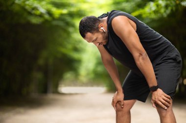 Exhausted handsome african american young guy jogging in public park, having break while getting ready for marathon, listening to music, using earbuds, panorama with copy space