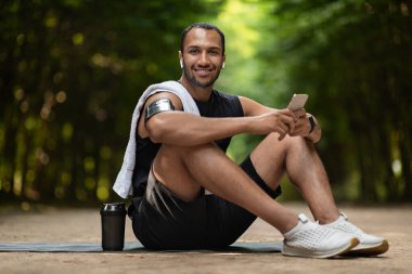 Cheerful attractive african american guy jogger sitting on yoga mat with towel on his shoulder, holding cell phone, using white earbuds, drinking protein cocktail, having break while training outdoors